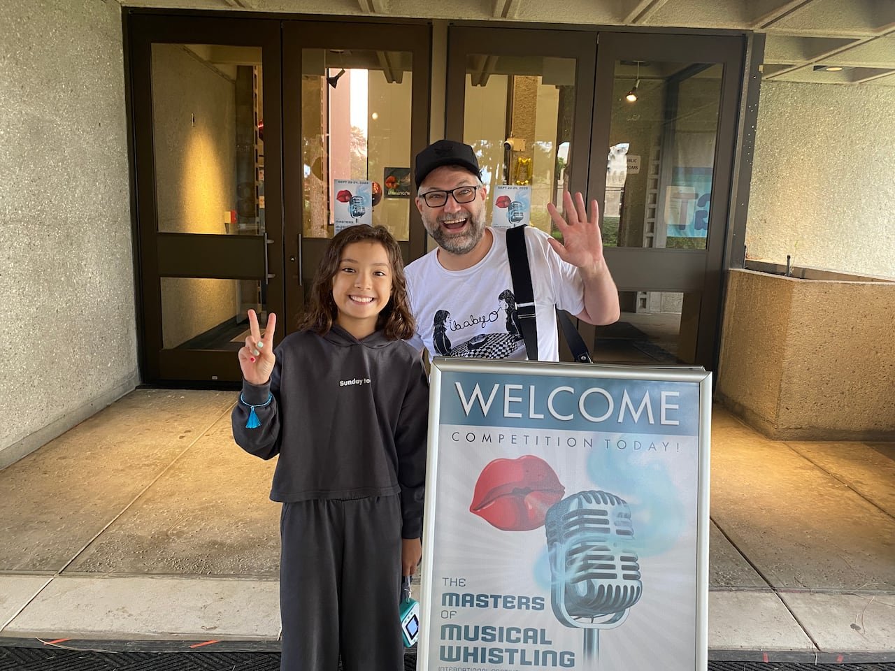 An adult man and young girl wave in front of a stone building's entrance. They're standing behind a sandwich board sign with the word "WELCOME" on it and a painting of an old fashioned microphone.