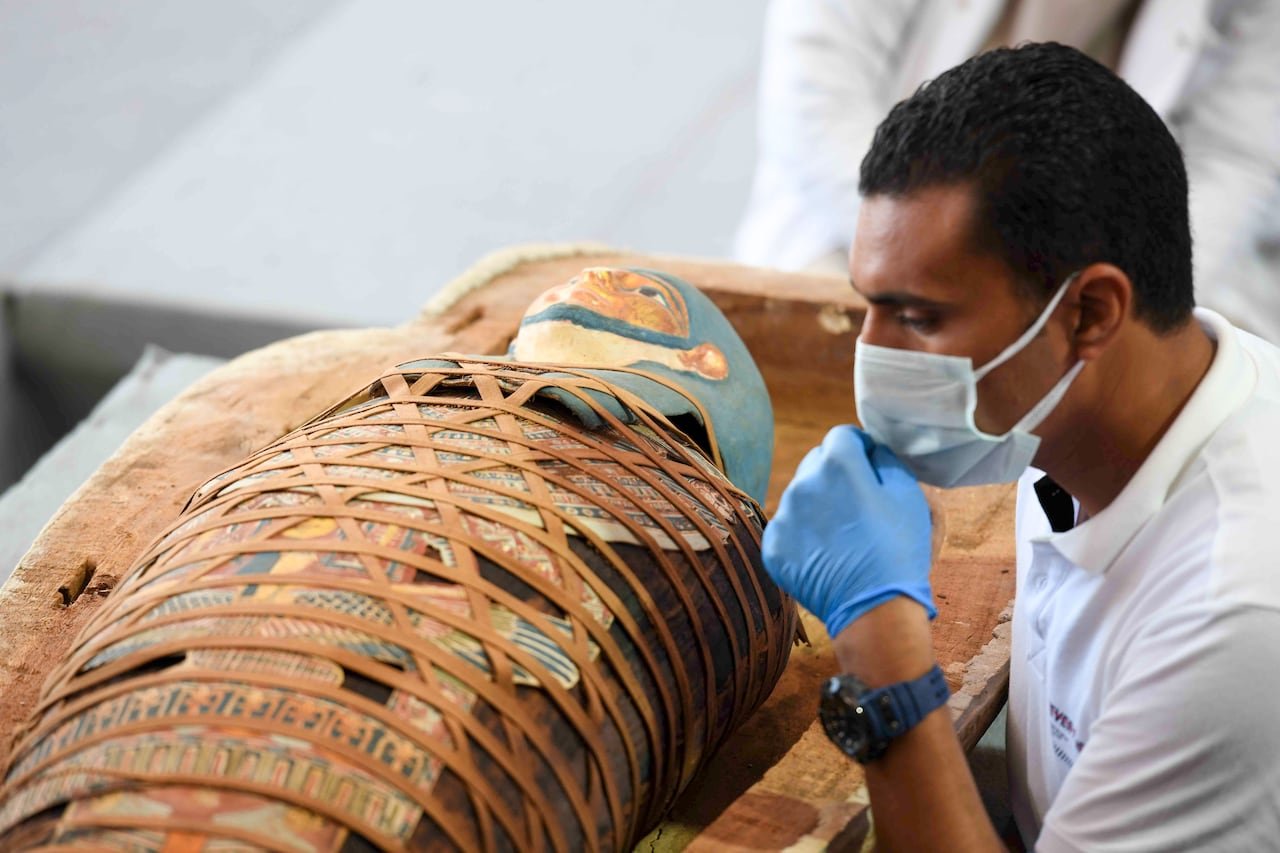 default-115 An archaeologist inspects an Egyptian mummy that is wrapped in an elaborate and colourful burial shroud. The archaeologist is wearing a medical mask and gloves.