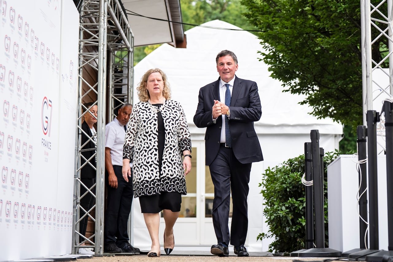 A man and a woman converse while walking outdoors. Surrounding them are signage and a white tent.