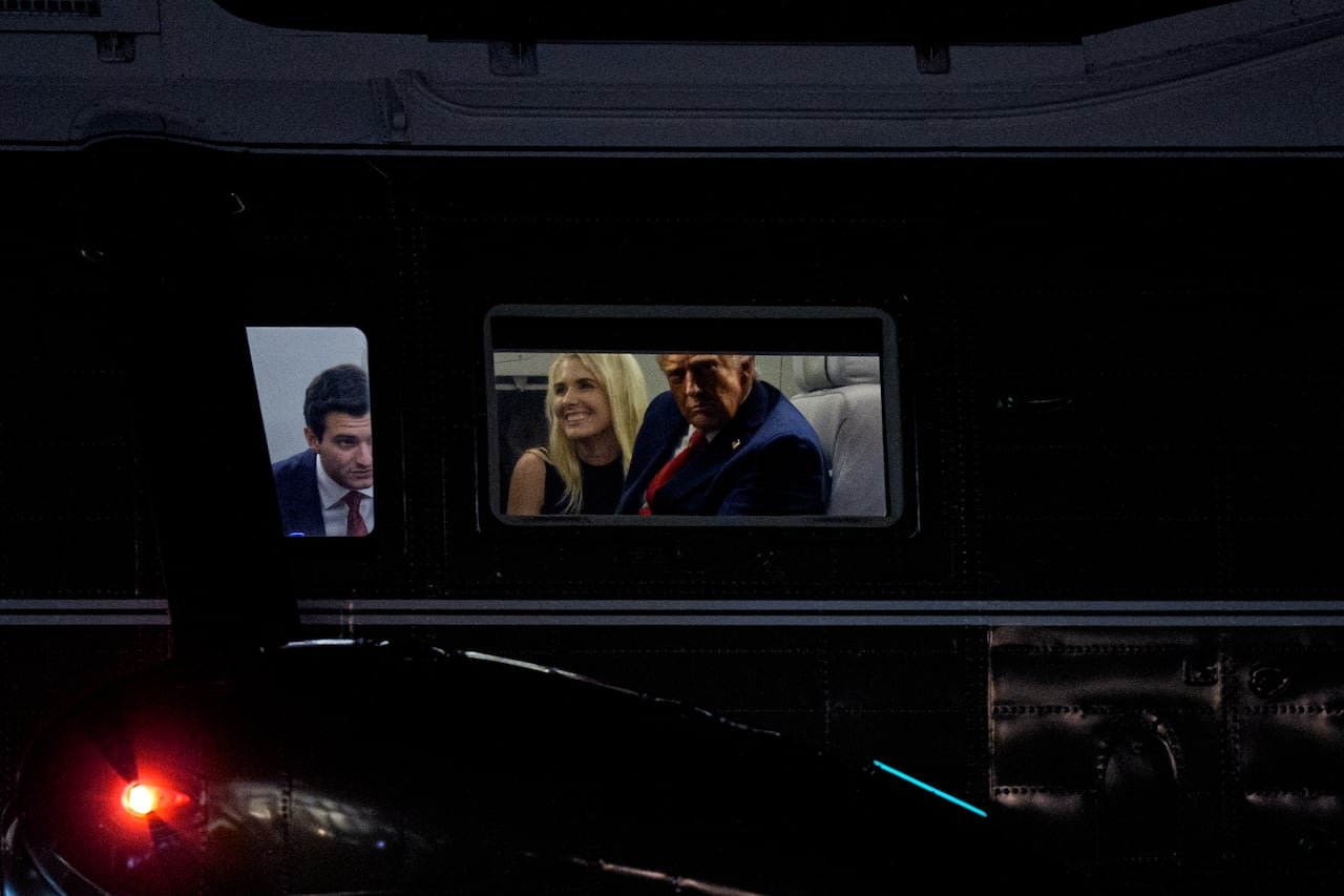 Two men in suits and a smiling woman with shoulder-length blonde hair are illuminated through a window, with darkness outside.