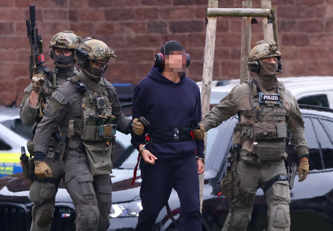 default-502 A man, with his face blurred, wearing headphones, a ballcap, and a hoodie, is seen being escorted by three police officers in camouflage uniforms with guns.