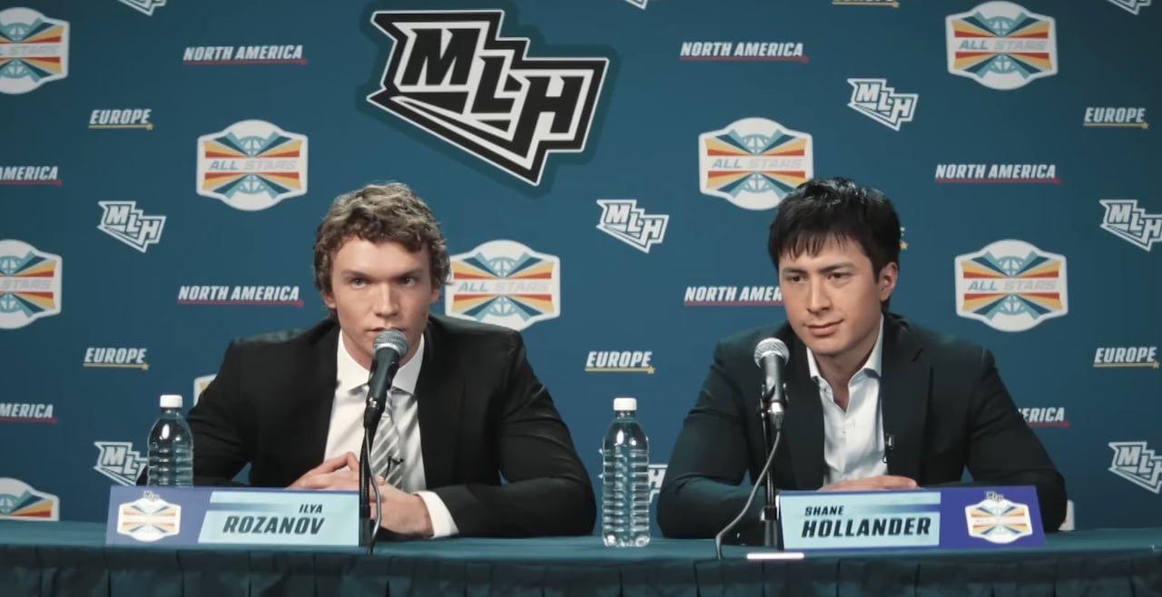 default-534 Two men sit down for a press conference. Both are wearing black blazers with white dress shirts underneath. Behind them is a series of logos